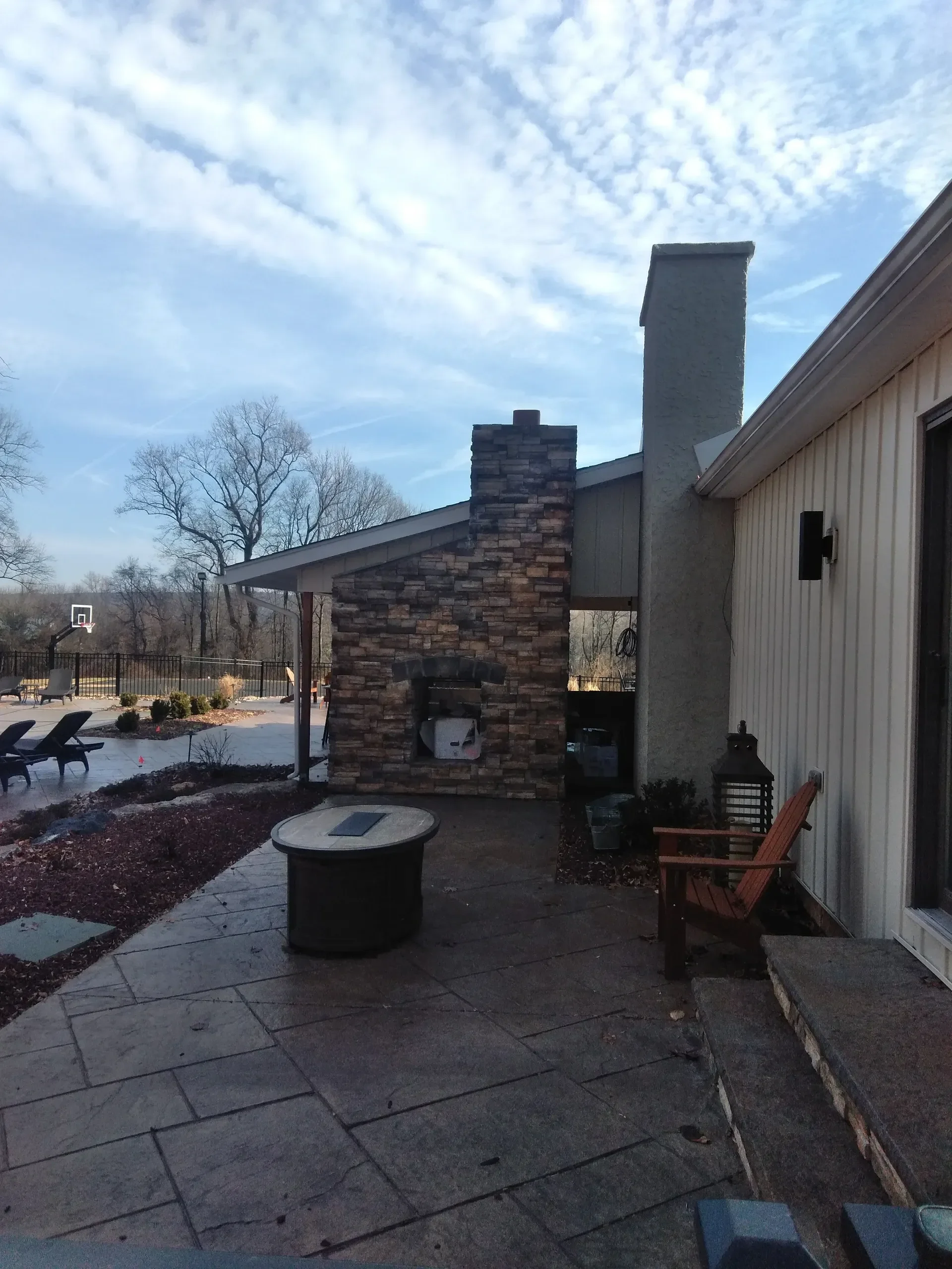 A view of a residential backyard patio featuring a stone veneer outdoor fireplace, a round fire pit, and stamped concrete paving under a cloudy sky.