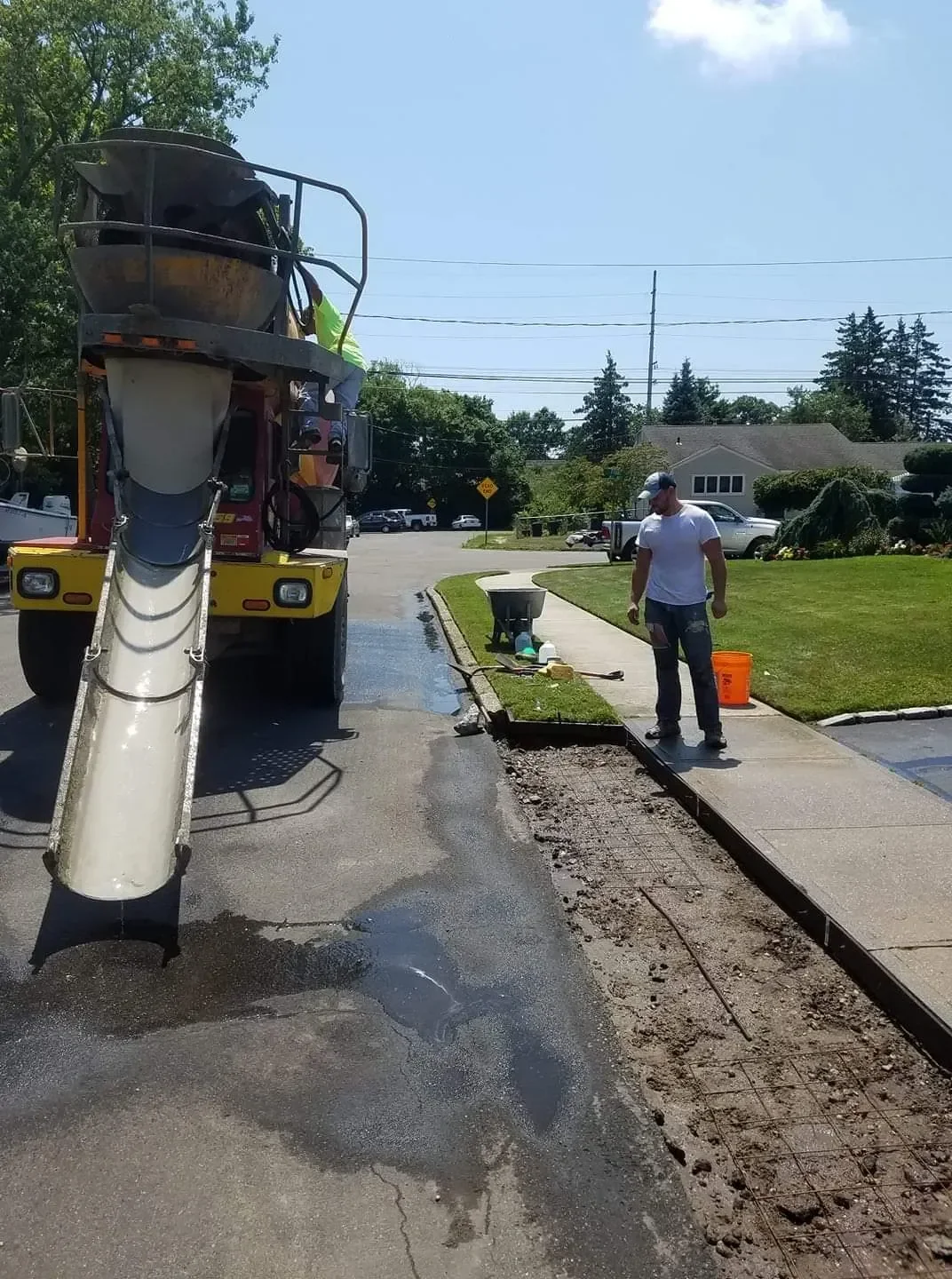 A cement mixer truck is positioned on a residential street next to workers preparing a trench with rebar mesh for pouring a new concrete curb and sidewalk section on a sunny day.
