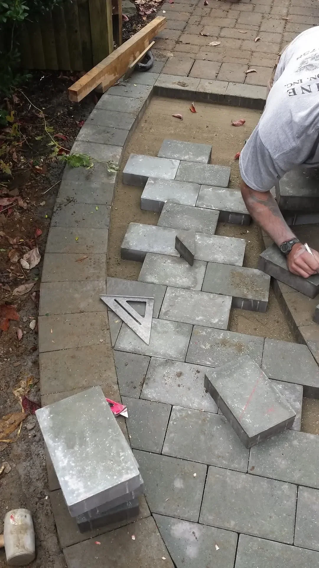 A worker installs gray rectangular pavers on a sand base, utilizing a speed square and marking stones near a newly constructed curved edge.