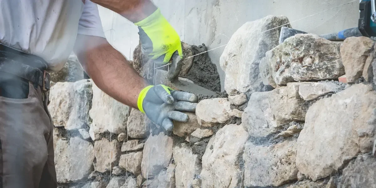 A construction worker wearing protective gloves uses a trowel to apply mortar between large, irregular stones while building a rustic wall.