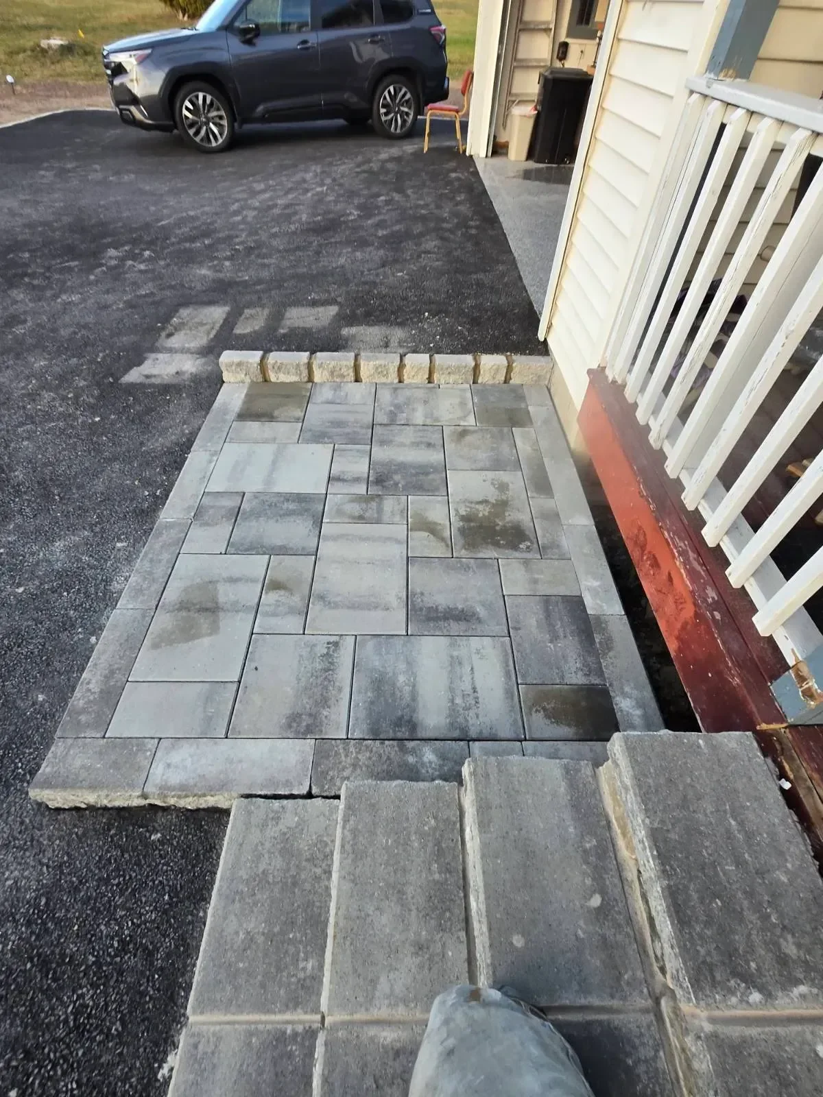 A newly installed gray paver patio landing and matching steps transition from the asphalt driveway to the house entrance, bordered by a small stone curb.