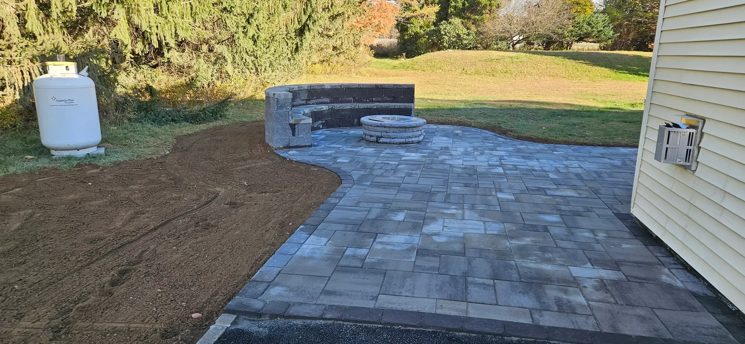 A newly installed gray paver patio featuring a curved stone seating wall and a circular fire pit is bordered by bare soil and a large white propane tank.