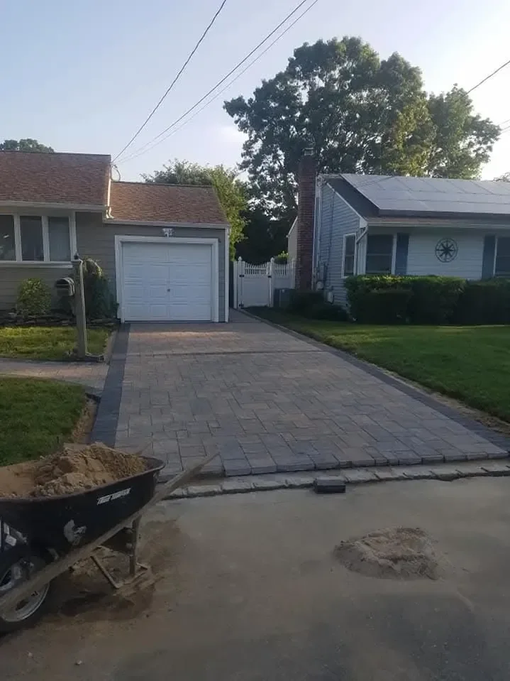 A newly laid interlocking paver driveway leads to a residential garage, with a wheelbarrow full of sand visible in the foreground indicating ongoing construction work.