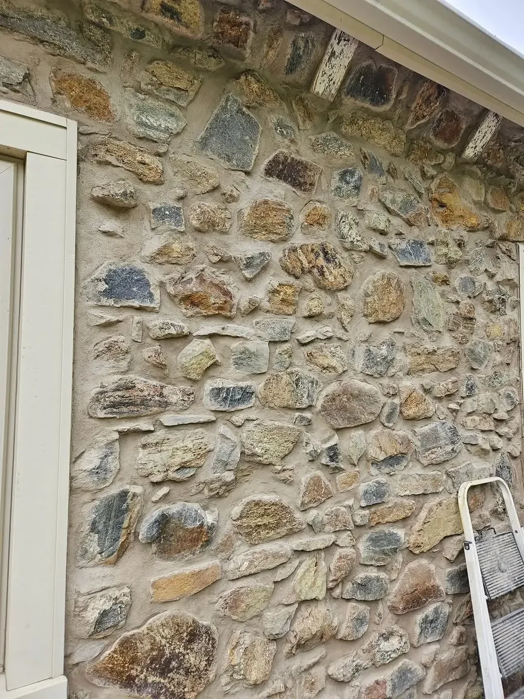 A close-up view shows an exterior wall constructed of multicolored natural stones set in thick mortar, featuring a window frame and a stepladder leaning against the surface.