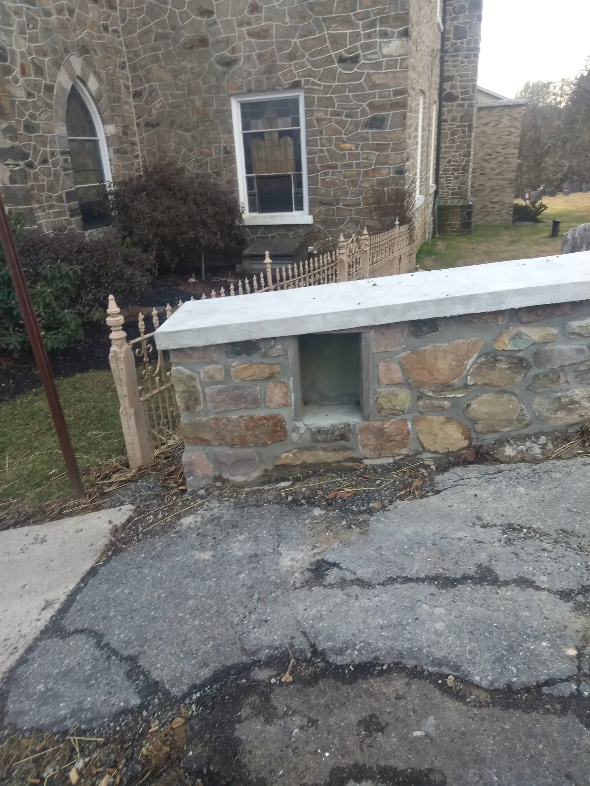 A newly capped fieldstone wall with a recessed niche stands before an older stone building featuring a gothic arched window and a decorative metal fence.